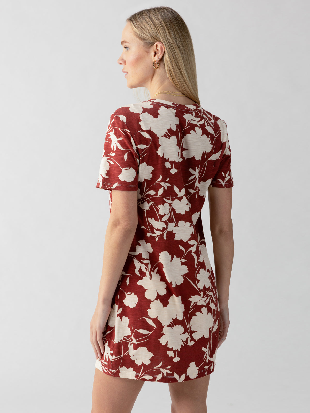 A woman with long blonde hair, standing and facing away from the camera, is dressed in the Sanctuary Clothing brand's short-sleeved "The Only One T Shirt Dress Warm Vista," featuring a vibrant red hue adorned with a charming white floral pattern. The backdrop is plain and light-colored.