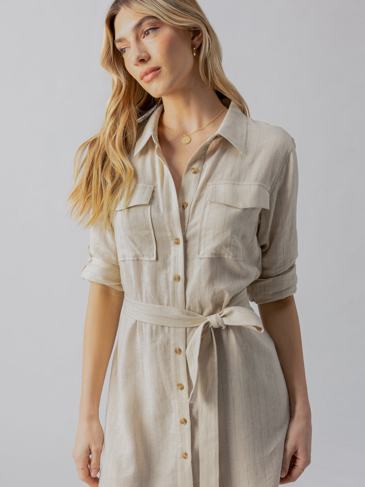 A woman with long blonde hair is wearing a Sanctuary Clothing Pocket Shirt Dress in Vineyard Stripe, featuring a belt tied at the waist. The dress includes two chest pockets, and the sleeves are rolled up. She is standing against a plain, light gray background.