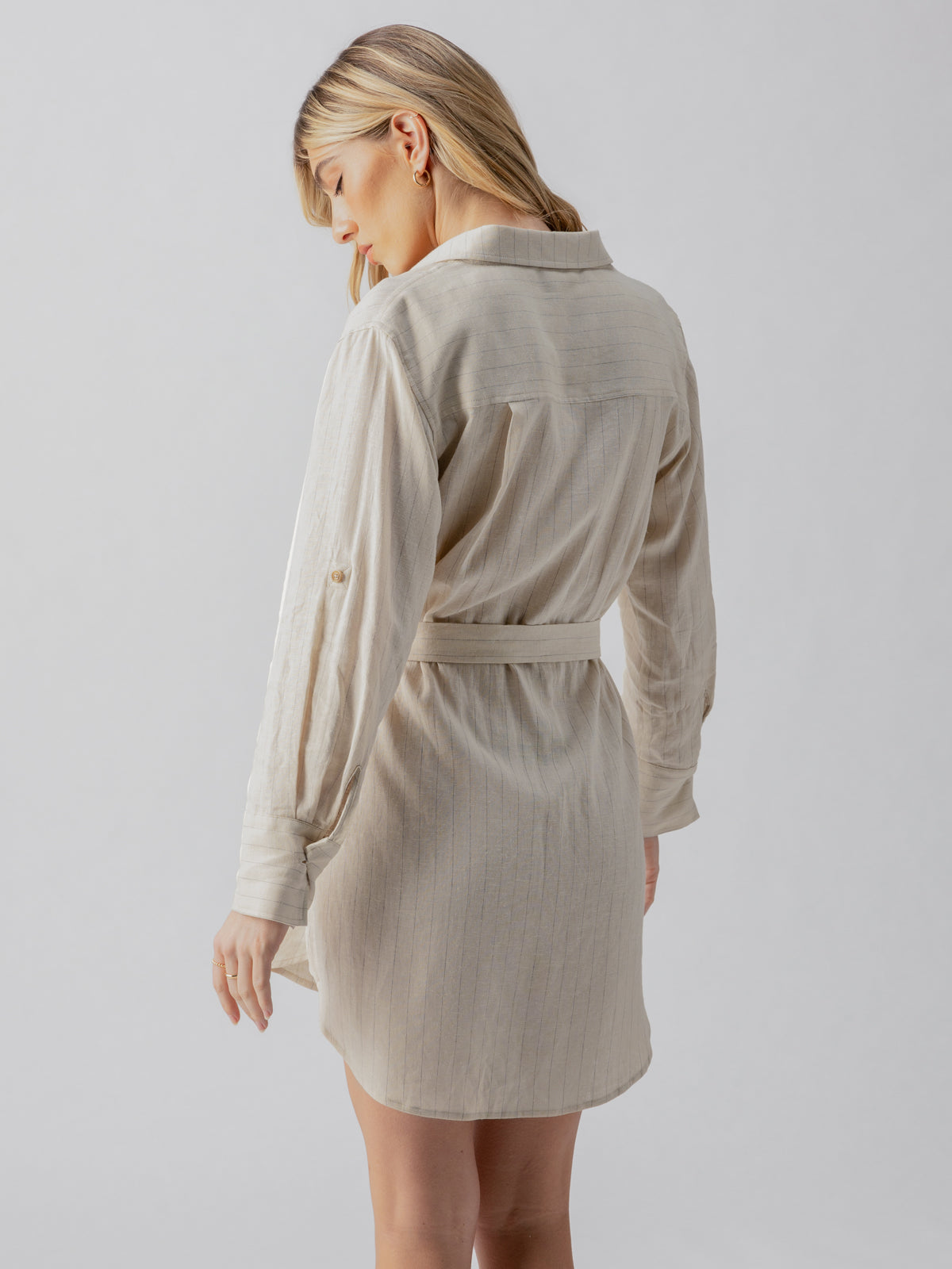 A woman with blonde hair is shown from the back, wearing the Pocket Shirt Dress Vineyard Stripe by Sanctuary Clothing. She stands against a light gray background, slightly turning her head to the side and looking downward, showcasing the dress's long sleeves, button-down design, and waist belt.