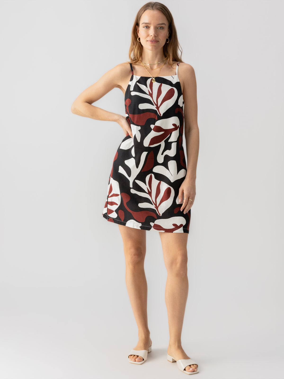 A woman stands confidently in a black, white, and red sleeveless Mini Shift Dress Mineral by Sanctuary Clothing, featuring bold, abstract leaf patterns. She rests one hand on her hip and pairs the dress with white sandals against a plain white background.