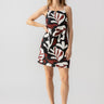 A woman stands confidently in a black, white, and red sleeveless Mini Shift Dress Mineral by Sanctuary Clothing, featuring bold, abstract leaf patterns. She rests one hand on her hip and pairs the dress with white sandals against a plain white background.