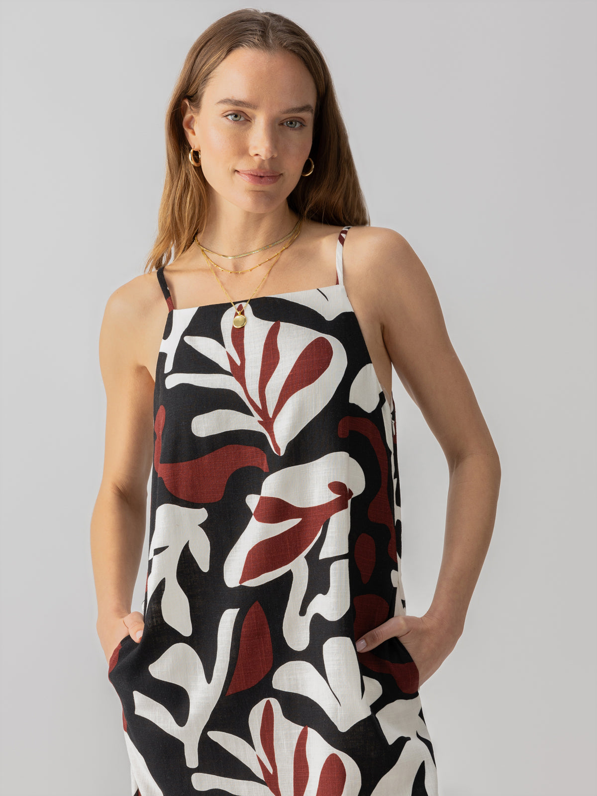 A person with long hair is wearing the "Mini Shift Dress Mineral" from Sanctuary Clothing. The dress features an abstract design in red, white, and black with thin straps. The person has their hands in the dress pockets and is looking directly at the camera with a slight smile. The background is plain and light-colored.