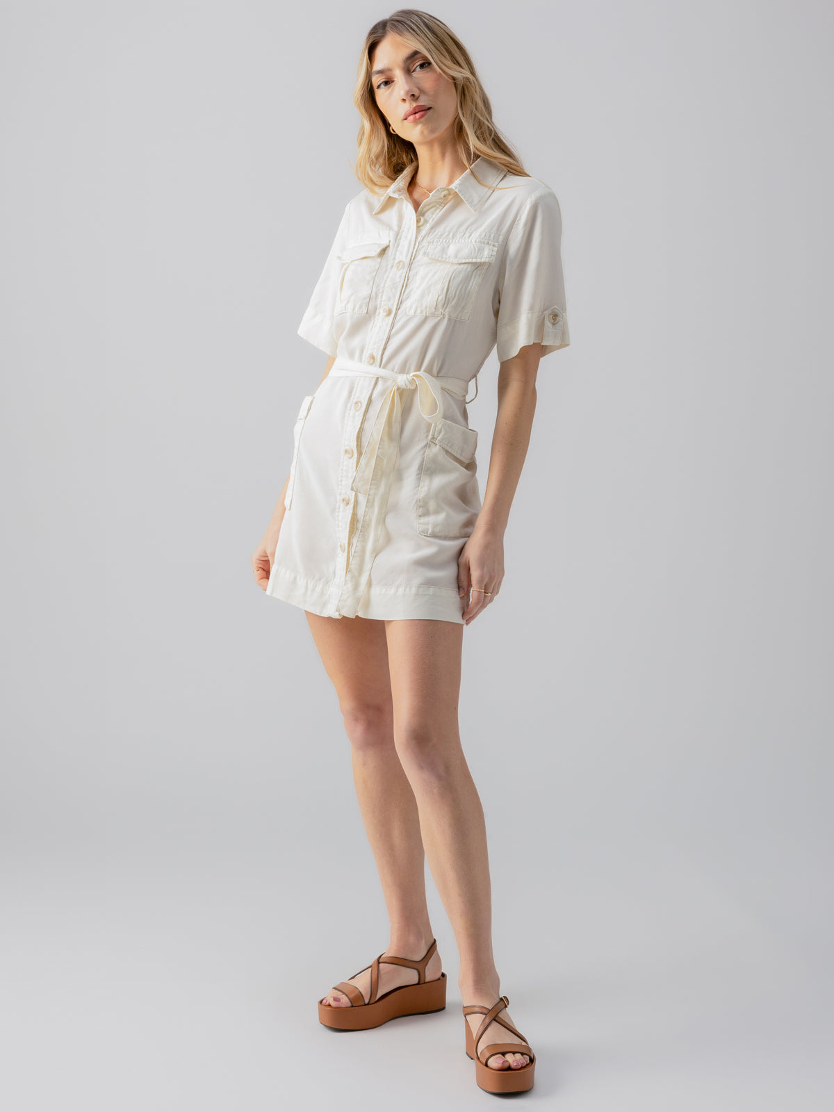 A woman stands against a plain background, wearing the Fluid Surplus Dress Birch by Sanctuary Clothing. The dress is a short-sleeve, button-down with pockets and a belt. She has long blonde hair and is looking at the camera, paired with brown wedge sandals.