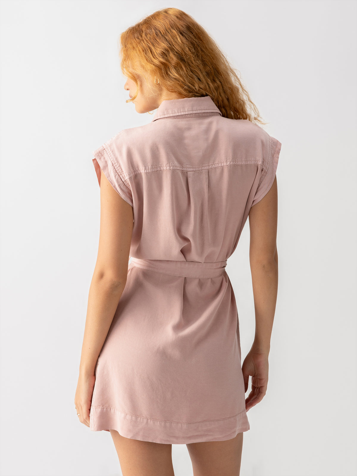 A person with long, wavy hair is seen from the back, wearing the Theo Surplus Dress Smokey Rose from Sanctuary Clothing. The short-sleeved dress features a light pink belted design with a loose fit and a collar, creating a casual yet stylish look. The background is plain white, emphasizing both the dress and the figure.