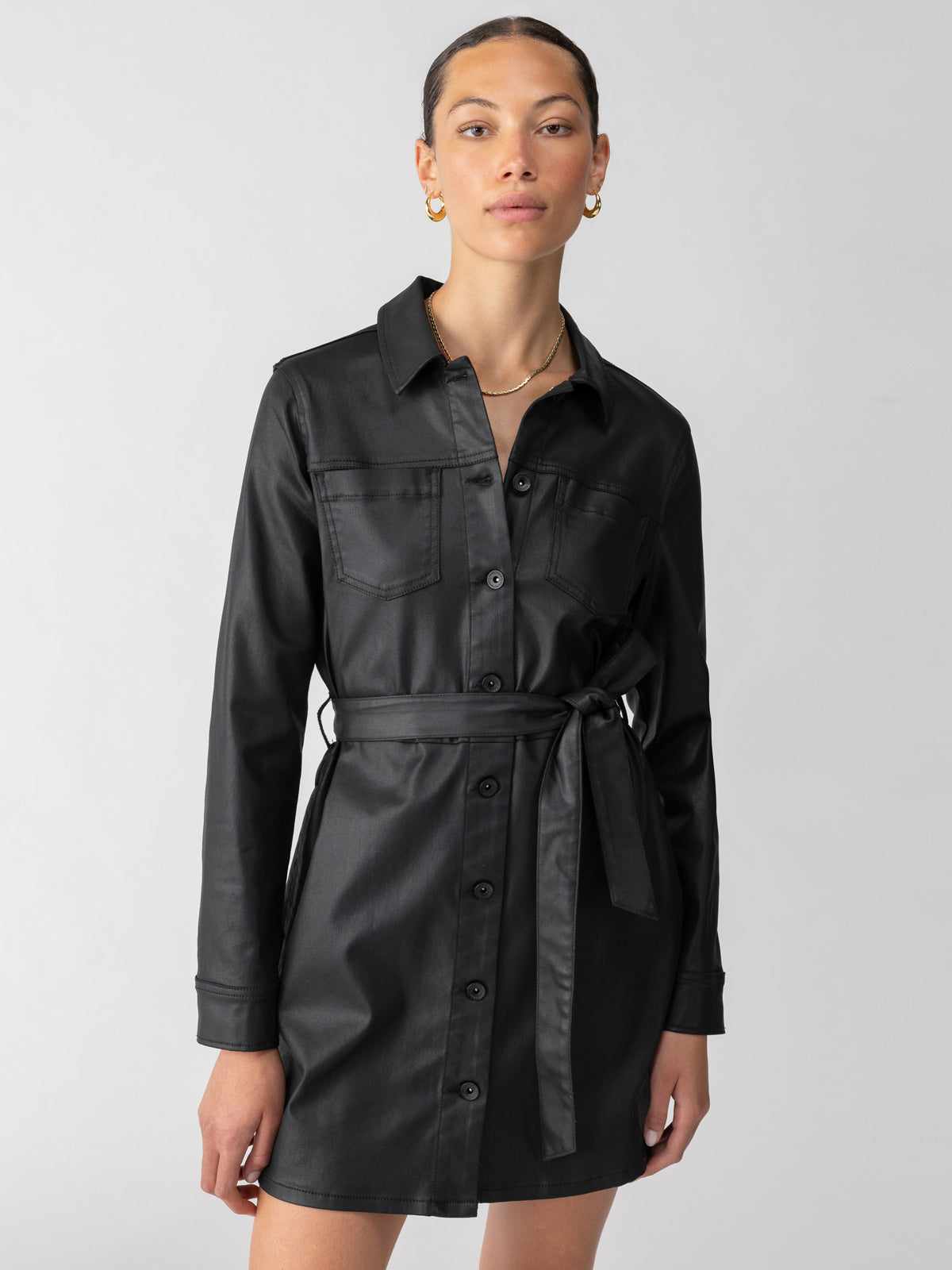 A person with short pulled-back hair wears the "Coated Denim Dress Black" by Sanctuary Clothing. This black, long-sleeve, button-up dress with a waist belt features two chest pockets. They are looking directly at the camera, standing against a plain, light-gray background.