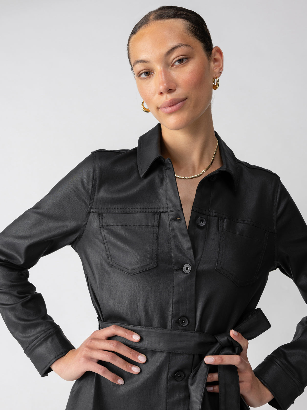 A woman with a confident expression is wearing a stylish Coated Denim Dress Black by Sanctuary Clothing, featuring a belted waist and button-down front. She accessorizes with gold hoop earrings and a thin gold necklace. Her hair is neatly pulled back, and she stands against a plain light grey background.