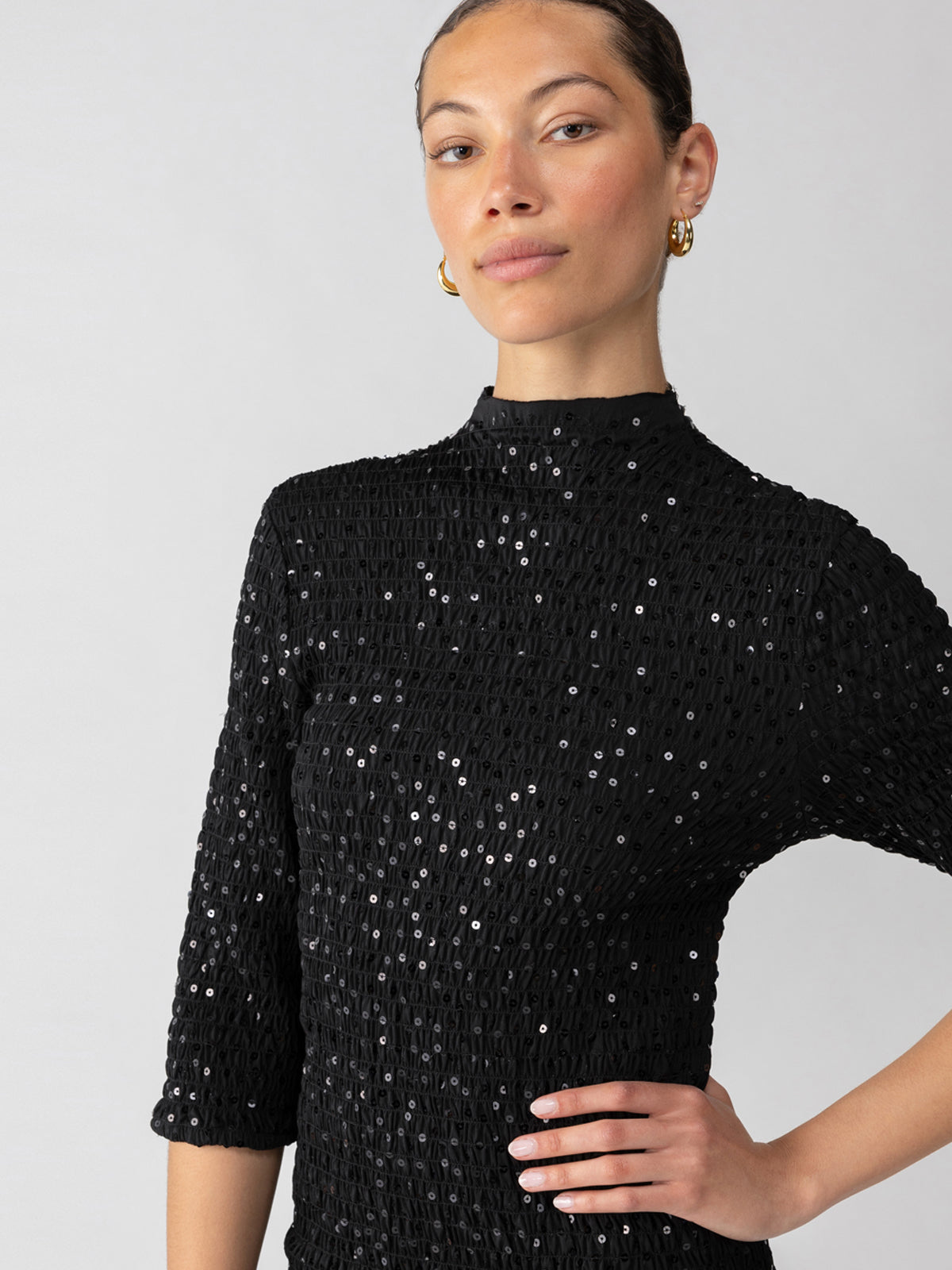 A person with a neutral expression is wearing the Holiday Dream Smocked Sequin Dress in black from Sanctuary Clothing. Their hair is pulled back, and they are accessorized with gold hoop earrings. The background is plain and light-colored.