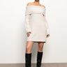 A woman stands confidently against a plain background, wearing the off shoulder dress in coconut smoke by Sanctuary Clothing and black knee-high boots. Her hair is styled neatly back, and she wears minimal jewelry.