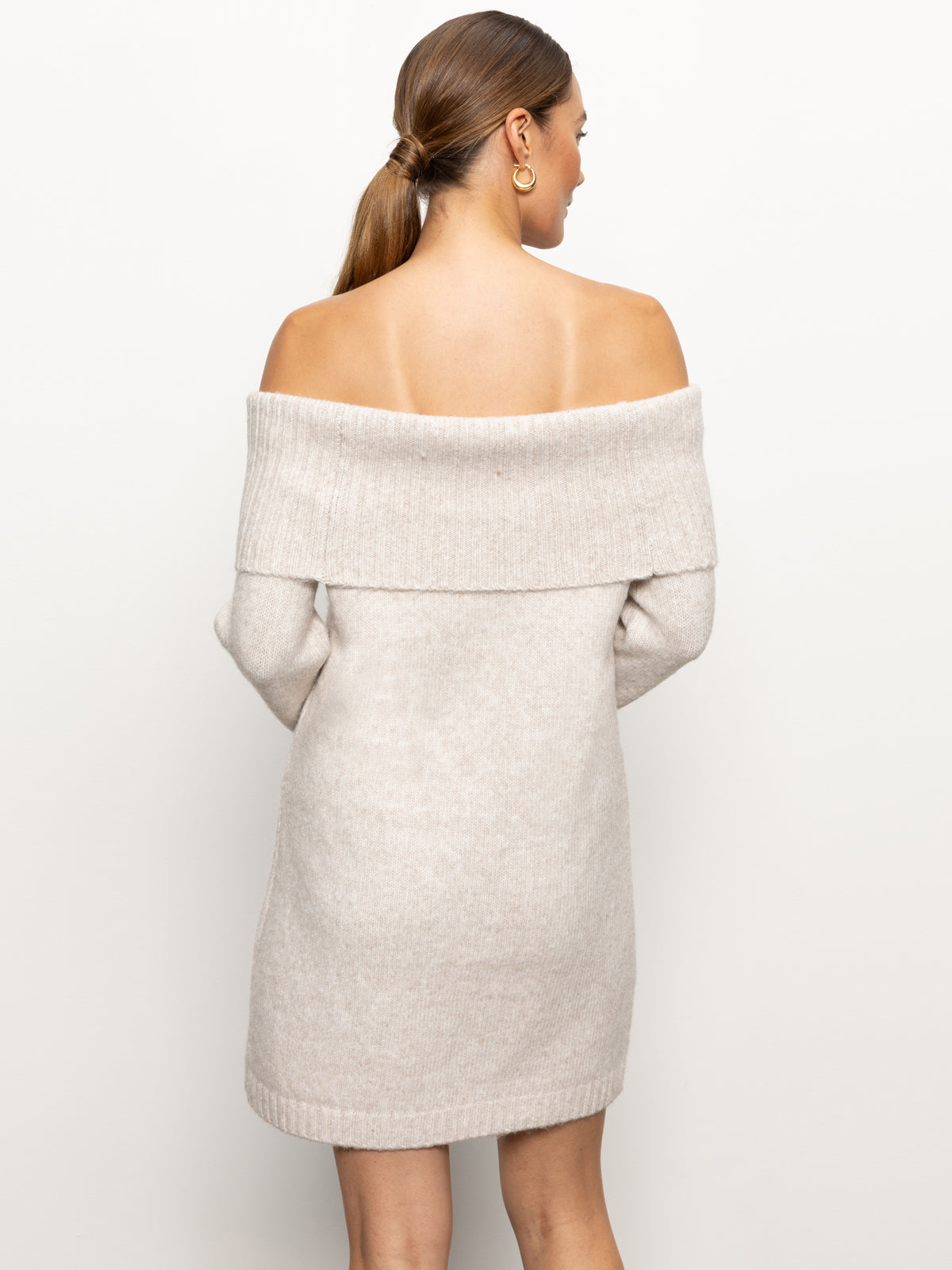 A woman with long hair in a ponytail is wearing Sanctuary Clothing's "Off Shoulder Dress Coconut Smoke." She is facing away from the camera, highlighting the soft, cream-knit texture of the dress against a plain background.