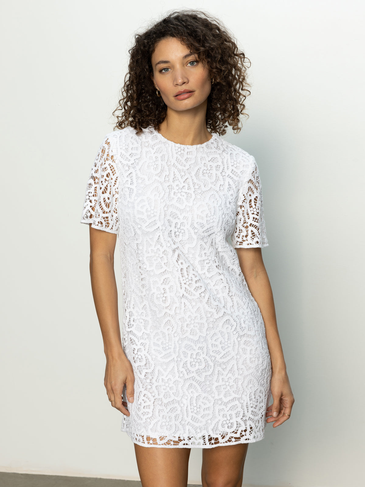 A woman in Sanctuary Clothing’s The Wanderer lace dress white stands against a light gray background. She has curly hair and gazes calmly at the camera.