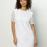 A woman in Sanctuary Clothing’s The Wanderer lace dress white stands against a light gray background. She has curly hair and gazes calmly at the camera.