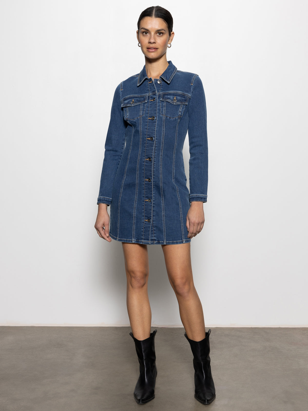 A woman stands against a white wall, wearing Sanctuary Clothing's sculpted denim mini dress royalty with long sleeves and a collar. She pairs it with black boots and her hair is pulled back.