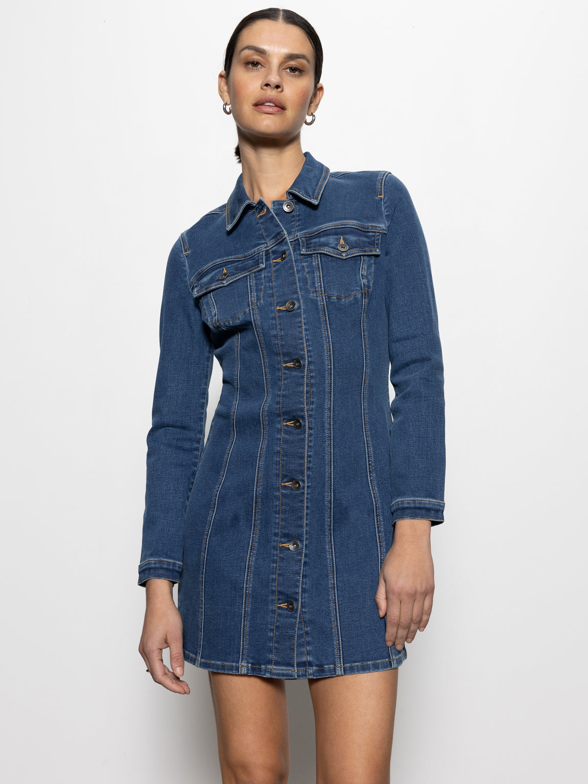 A woman models the Sanctuary Clothing "sculpted denim mini dress royalty," featuring long sleeves and button detailing, against a plain white background.