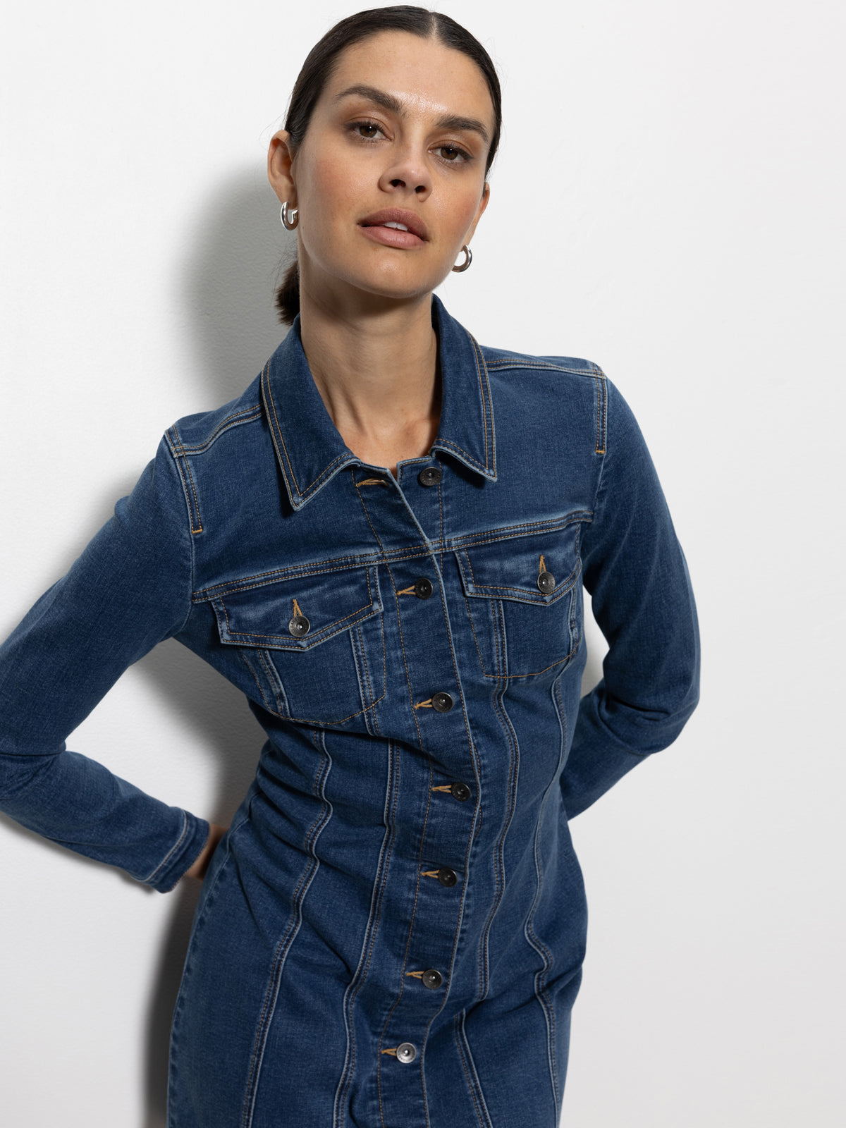 A person in Sanctuary Clothing's sculpted denim mini dress royalty stands against a white wall, with their dark hair tied back and small hoop earrings. They have a neutral expression and slightly placed arms at their back.