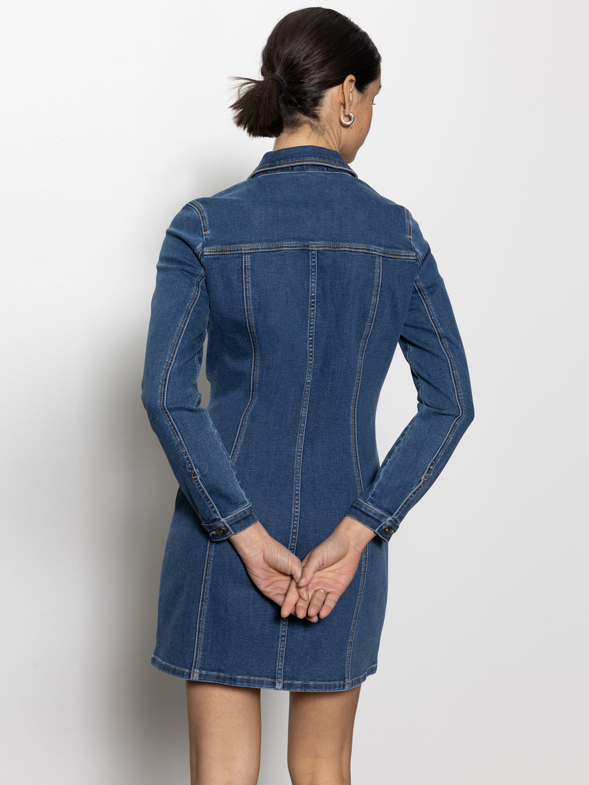 A person with dark hair in a bun, wearing the "sculpted denim mini dress royalty" by Sanctuary Clothing, stands facing away with their hands clasped behind their back against a plain, light-colored background.