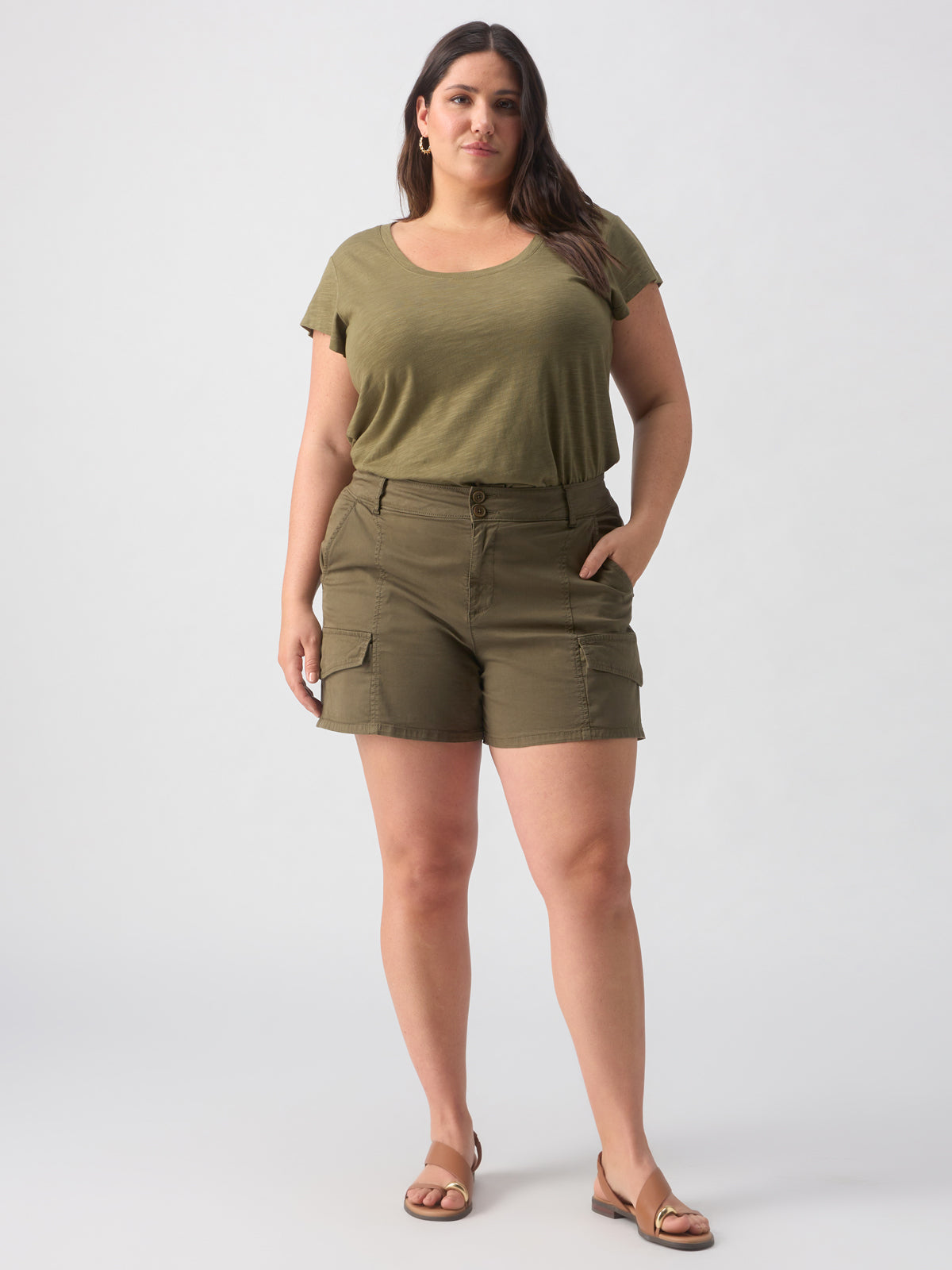 A woman stands against a plain background wearing Sanctuary Clothing's Rebel Short Hiker Green Inclusive Collection, with green short-sleeve top, olive green cargo shorts featuring pockets, and tan sandals. She has long dark hair, some of which is tucked behind her ear, and she maintains a neutral expression with her hands in her pockets.