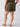 A person is shown from the back wearing Sanctuary Clothing's Rebel Short Hiker Green Inclusive Collection, featuring utility pockets. They are paired with a dark green top and tan low-heeled sandals, set against a plain, light-colored backdrop.