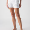 A person stands facing forward, wearing Sanctuary Clothing's Rebel Short in Brilliant White, which features two buttons and side pockets, paired with tan sandals. The background is plain and light-colored.