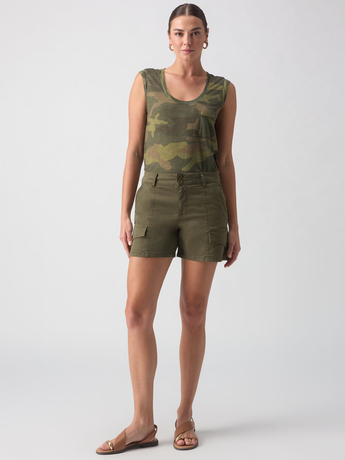 A person stands against a plain background, wearing Sanctuary Clothing's Rebel Short Hiker Green camouflage sleeveless top, olive green cargo shorts, and brown sandals. The individual has their hair pulled back and is looking directly at the camera.