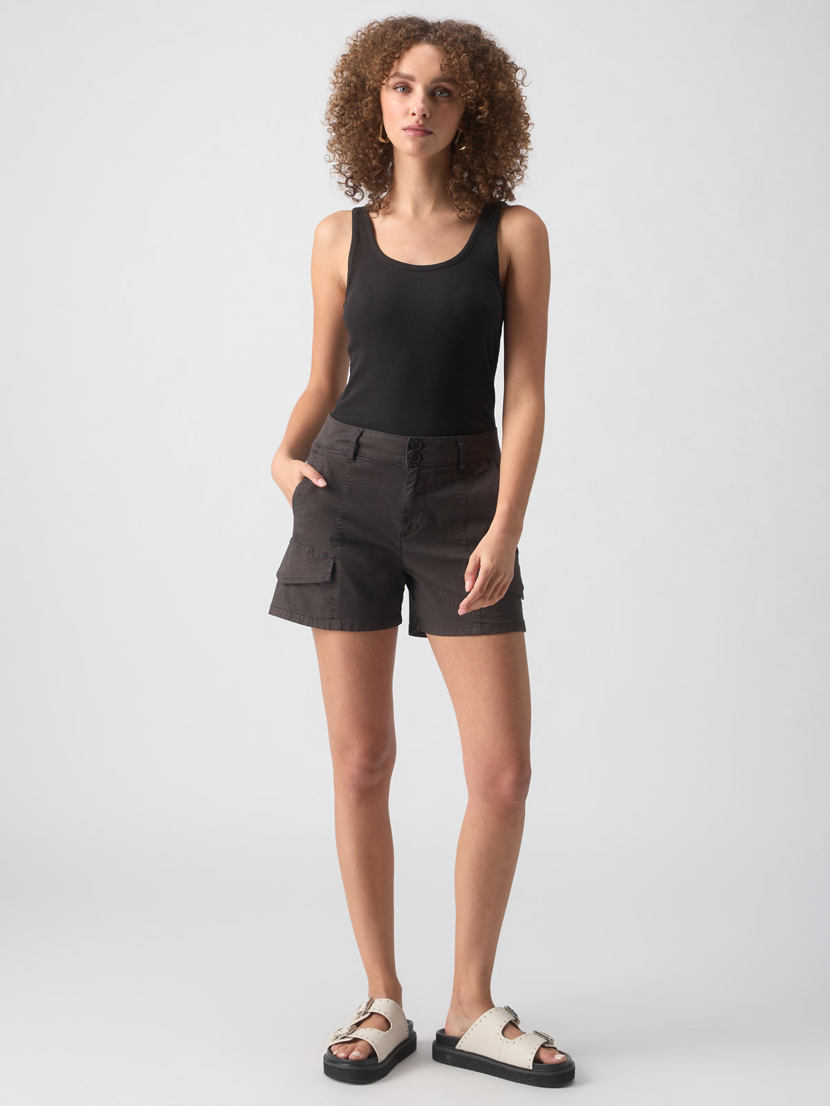 A person with curly hair, wearing a black sleeveless top, Sanctuary Clothing's Rebel Short in Obsidian, and white sandals stands against a plain background. They have their left hand in their pocket and appear relaxed.