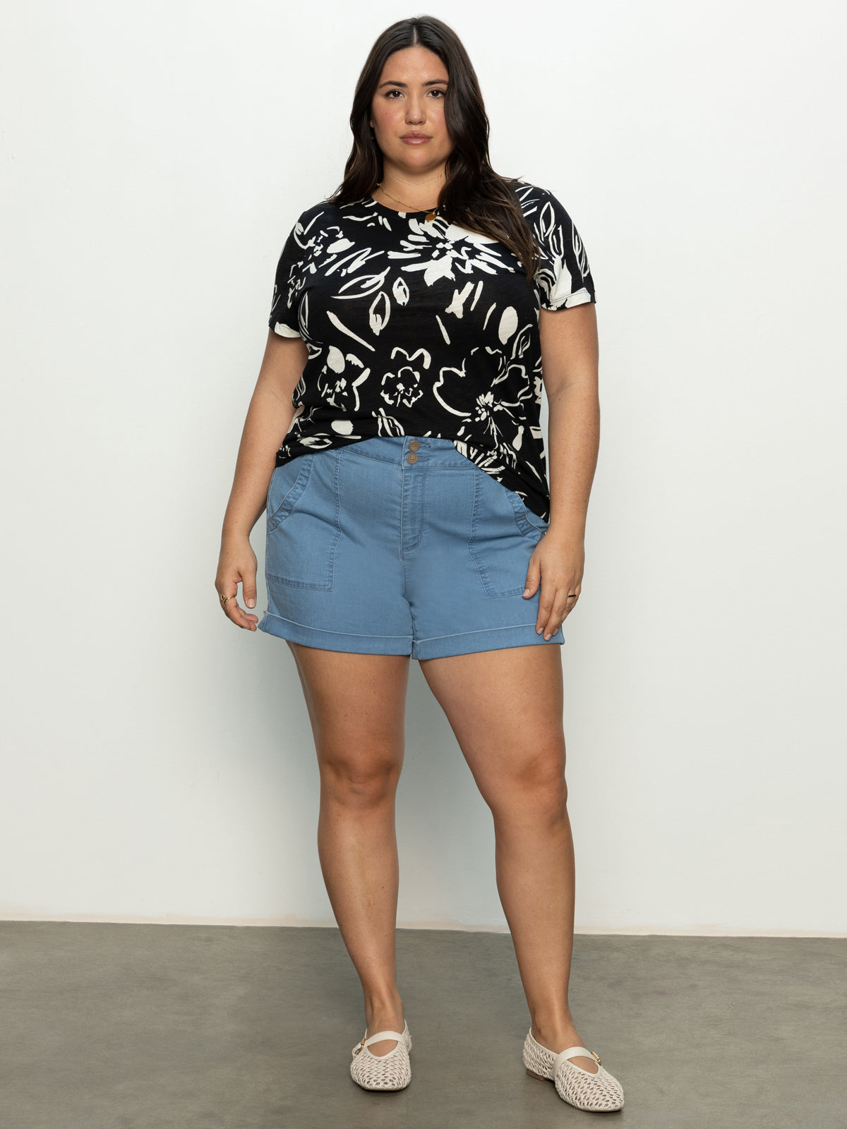 A woman wearing Sanctuary Clothing's renegade shorts from the tides inclusive collection, paired with a black and white floral shirt and beige shoes, stands against a plain background.