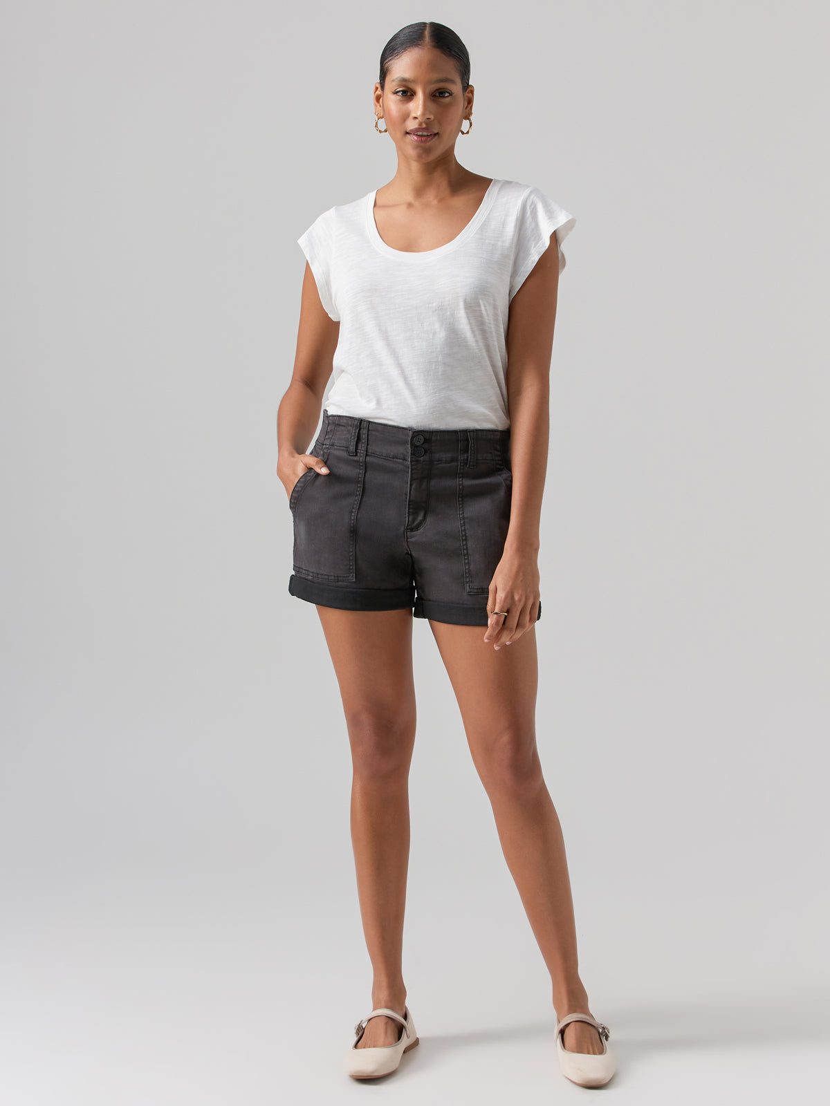 A person stands confidently against a plain background, wearing a white, short-sleeved top and the Renegade Short Black from Sanctuary Clothing. They have their hands in their pockets and are wearing beige ballet flats. The person is accessorized with hoop earrings.