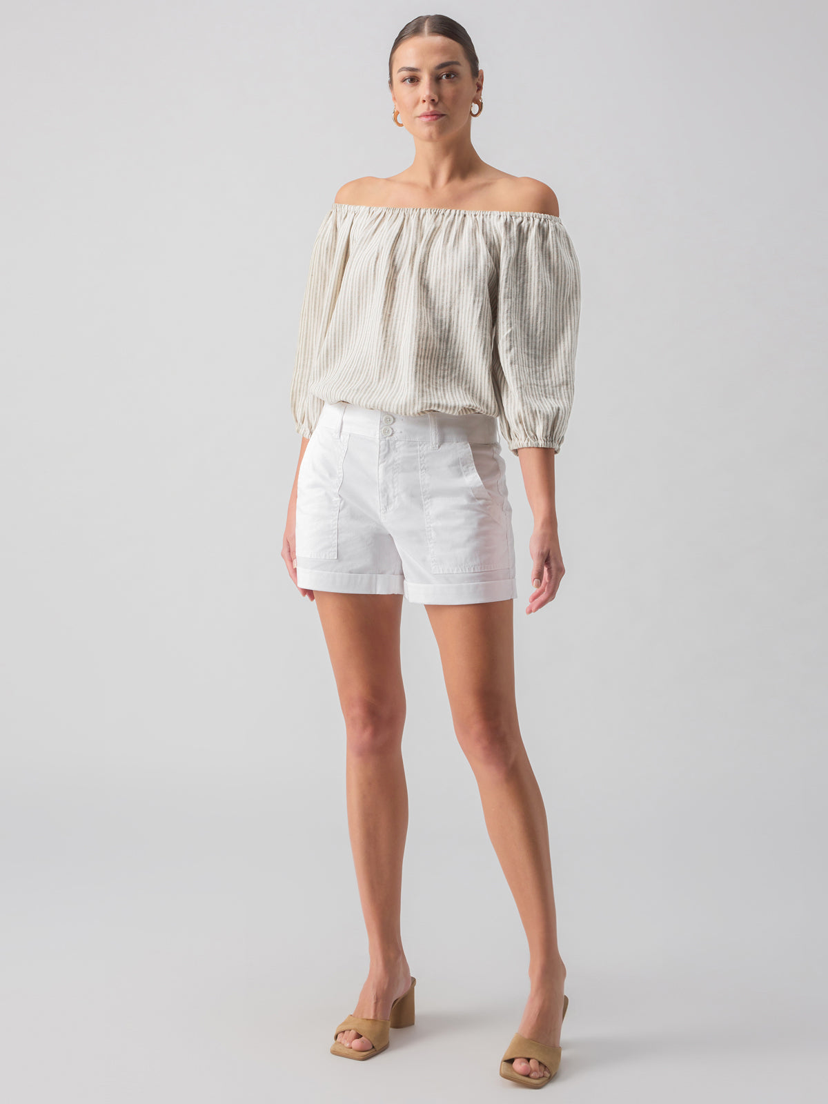 A person stands against a plain background wearing an off-shoulder, beige textured blouse and the Renegade Short White from Sanctuary Clothing. They have light-colored sandals with a low heel and hoop earrings. Their hair is pulled back.