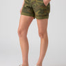 A person is wearing the Sanctuary Clothing Renegade Short Renew Camo with hands in the pockets, a tan-colored top, and beige sandals with black soles. The image focuses on the outfit from the waist down, set against a plain light gray background.