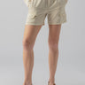 A person is dressed in the Relaxed Rebel Short Birch from Sanctuary Clothing, paired with a cream-colored knitted sweater and brown sandals. With their hands tucked into the shorts' pockets, they stand against a plain gray background, their face not visible.