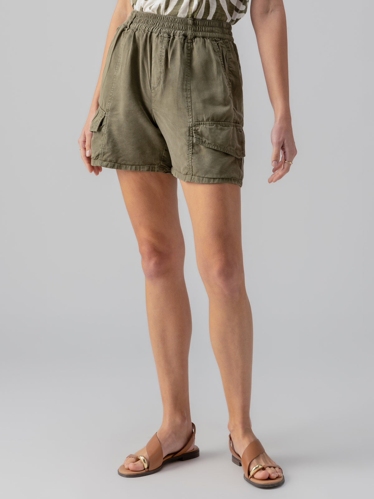 A person wearing the Sanctuary Clothing Relaxed Rebel Short in Burnt Olive and a white striped top stands against a plain background. The shorts feature multiple pockets, and the person is wearing brown sandals. Their hands are relaxed by their sides.