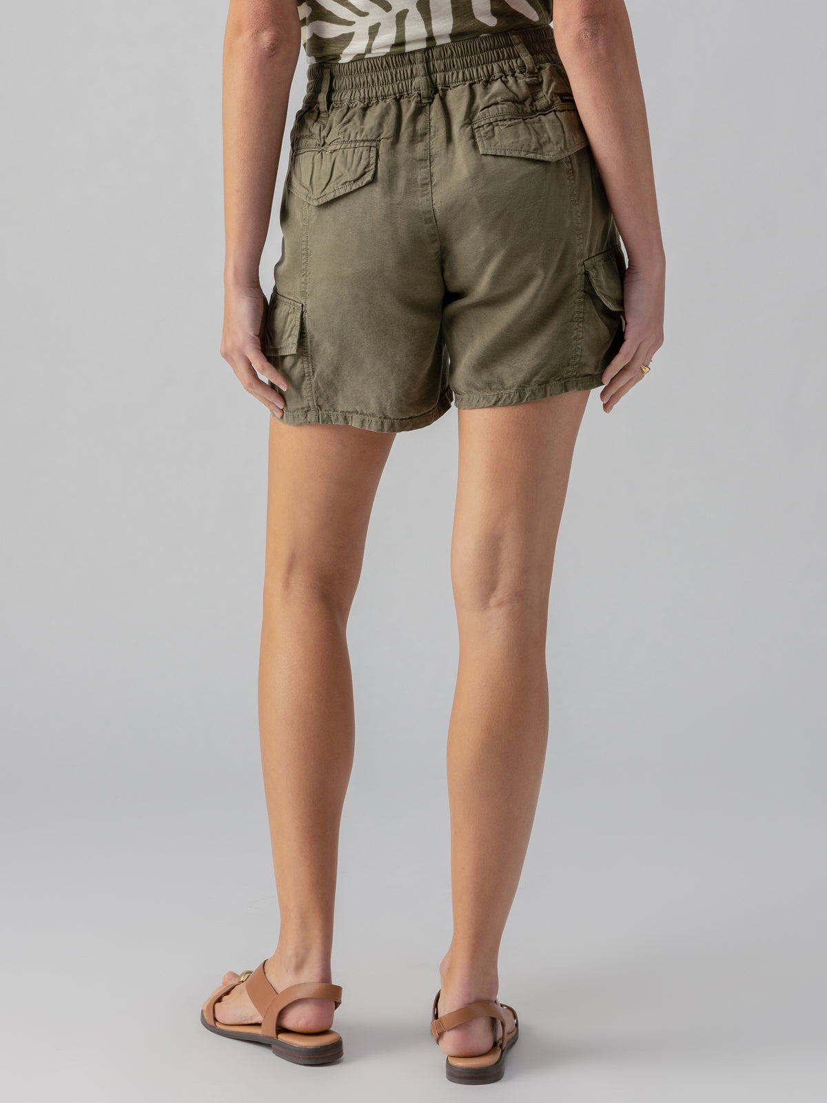 A person stands facing away from the camera, wearing Sanctuary Clothing's Relaxed Rebel Short in Burnt Olive paired with a beige patterned top. They are wearing brown sandals and have a ring on their right hand. The background is plain and grey.