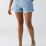 A person stands against a plain white background wearing the Beachy Cuffed Short in Pale Blue from Sanctuary Clothing, paired with a white top tucked in and white flats. Their hands are casually resting in the pockets of the shorts, creating a relaxed and summery look.