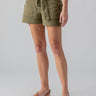 A person is wearing the Reissue Sash Short in Burnt Olive from Sanctuary Clothing, paired with a white tucked-in top. The person is also wearing tan flat sandals with a simple strap design. The background is plain light gray.