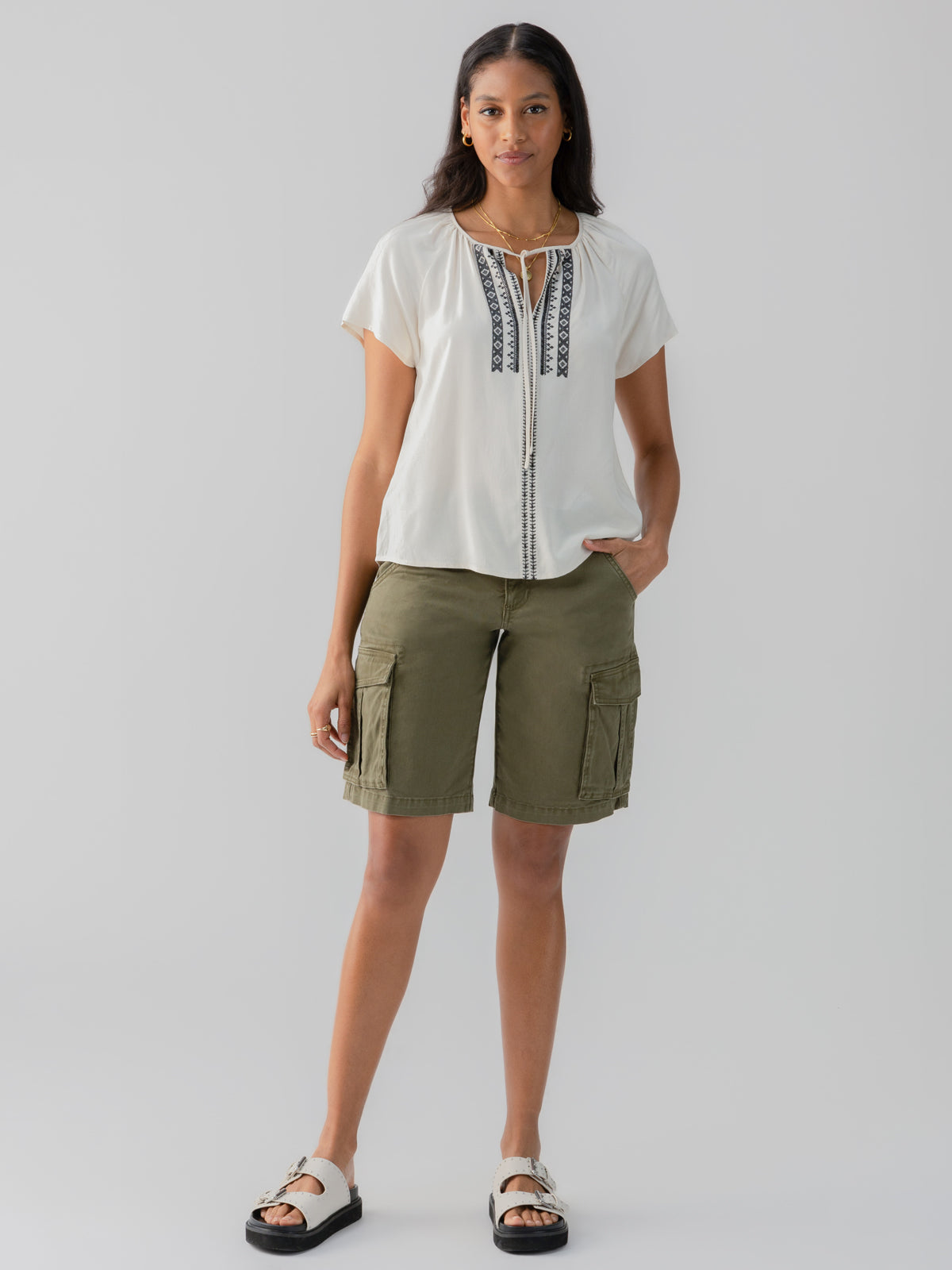 A woman stands against a plain background, dressed in an ivory blouse adorned with dark embellishments along the neckline and Sanctuary Clothing's Cargo Long Short Burnt Olive. She pairs her outfit with light-colored sandals. One hand rests in a pocket of her shorts as she looks forward.