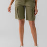 A person wearing the Cargo Long Short Burnt Olive by Sanctuary Clothing and a white top stands against a plain background. The shorts feature multiple pockets, and the person is wearing white sandals with black soles. The person's arms and upper legs are visible.
