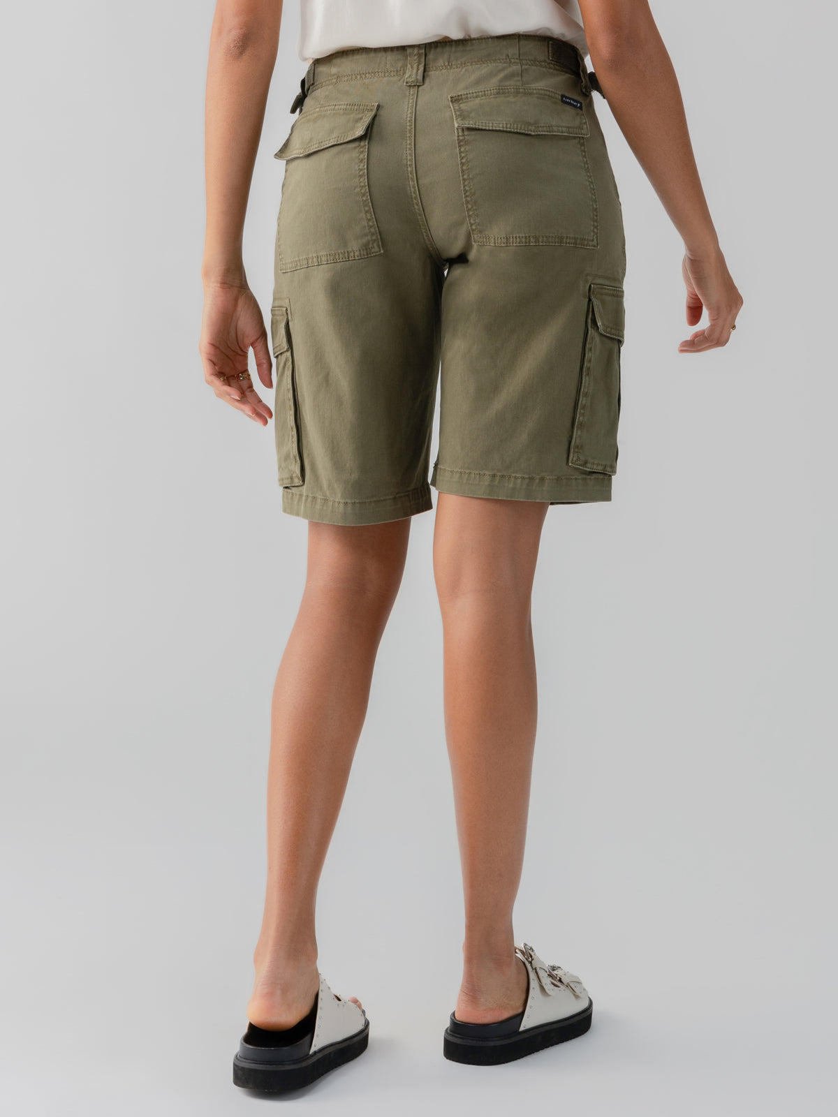 A person is shown from the back, wearing a white top and Sanctuary Clothing's Cargo Long Short in Burnt Olive, featuring multiple pockets. The individual is also wearing white slip-on shoes with black soles, against a plain light gray background.