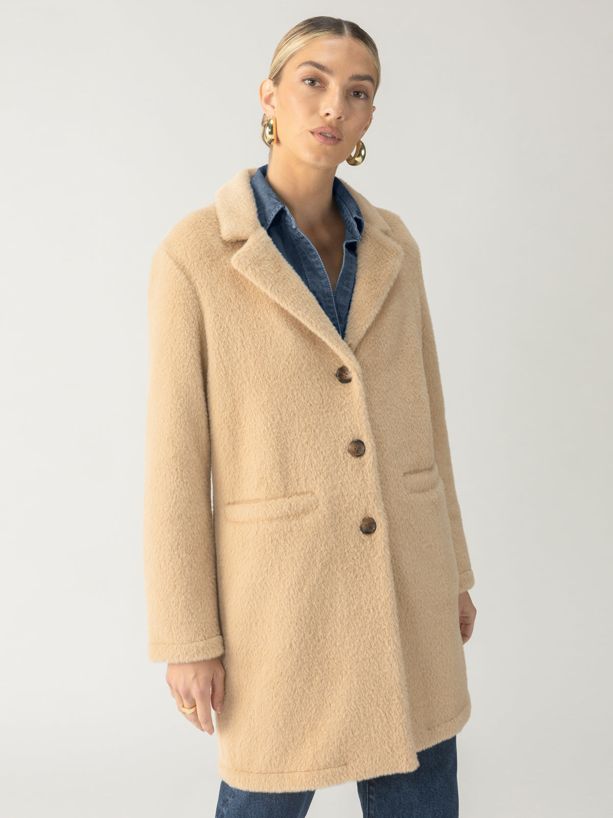 A person stands against a plain background wearing the Hometown Jacket Camel by Sanctuary Clothing, characterized by its beige wool texture, three buttons, and notch lapels. They have slicked-back hair and gold hoop earrings. Underneath the coat, they are wearing a denim shirt. Their facial expression is neutral.