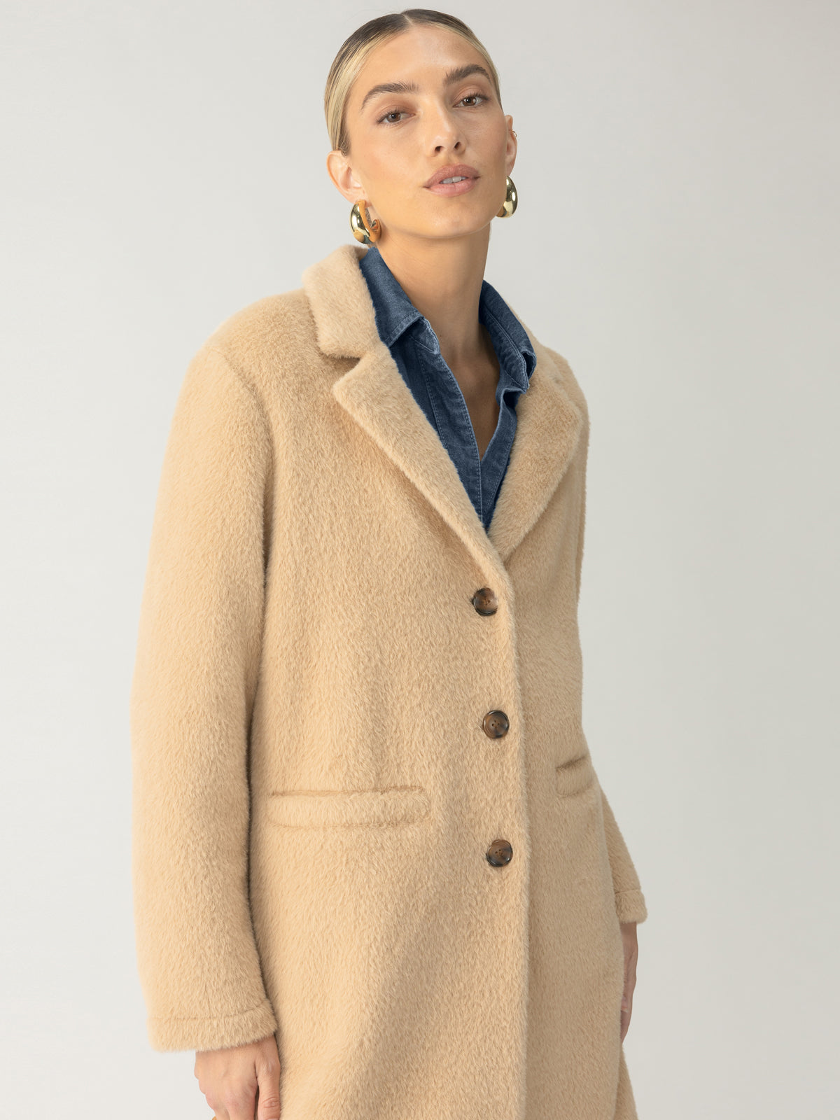 A person with slicked-back hair wears the knee-length, wool-textured Hometown Jacket Camel from Sanctuary Clothing, featuring large brown buttons. They pair it with a blue collared shirt and gold hoop earrings while posing against a plain, light-colored background.