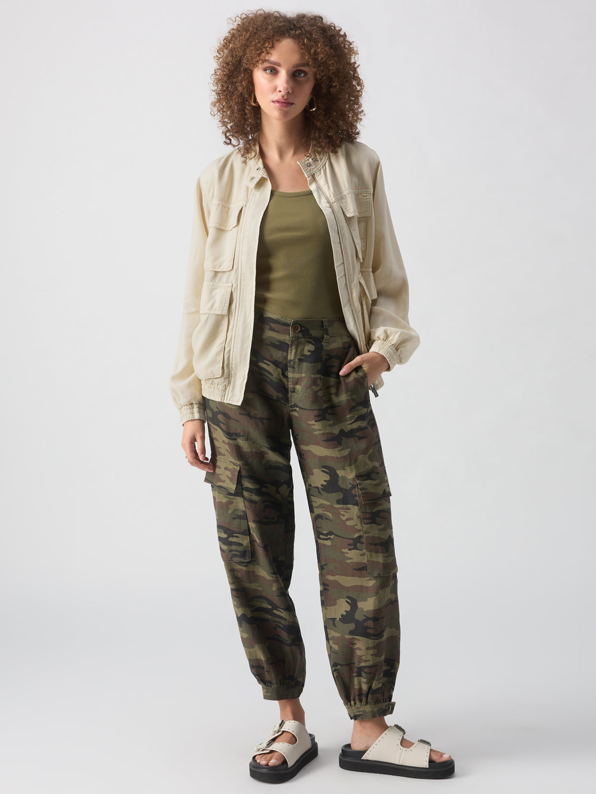 A person with curly hair wears the Field Utility Jacket Birch by Sanctuary Clothing over an olive green top, paired with camouflage cargo pants and cream-colored sandals with black socks. They have one hand in their pocket and stand against a neutral background.