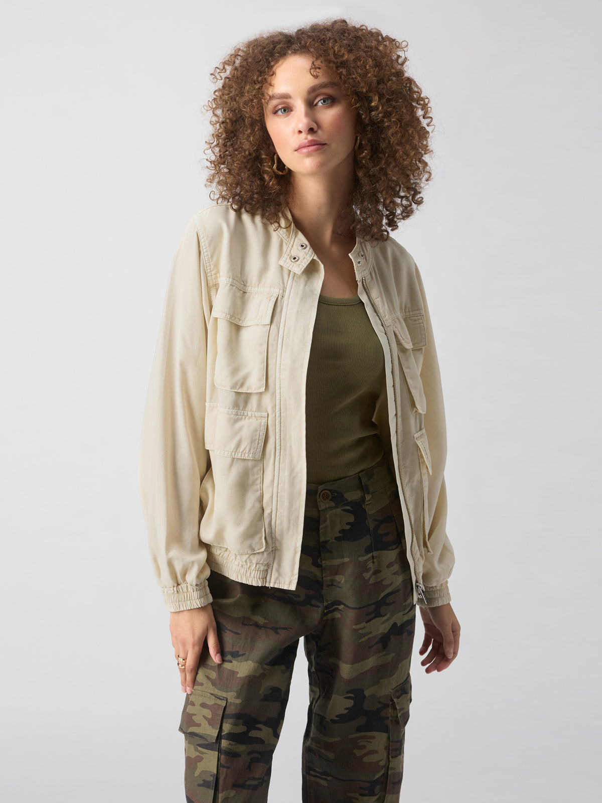 A person with curly hair stands against a neutral background, wearing the Field Utility Jacket Birch from Sanctuary Clothing over a green top and camouflage cargo pants. Their hands are relaxed at their sides, and they have a calm expression on their face.