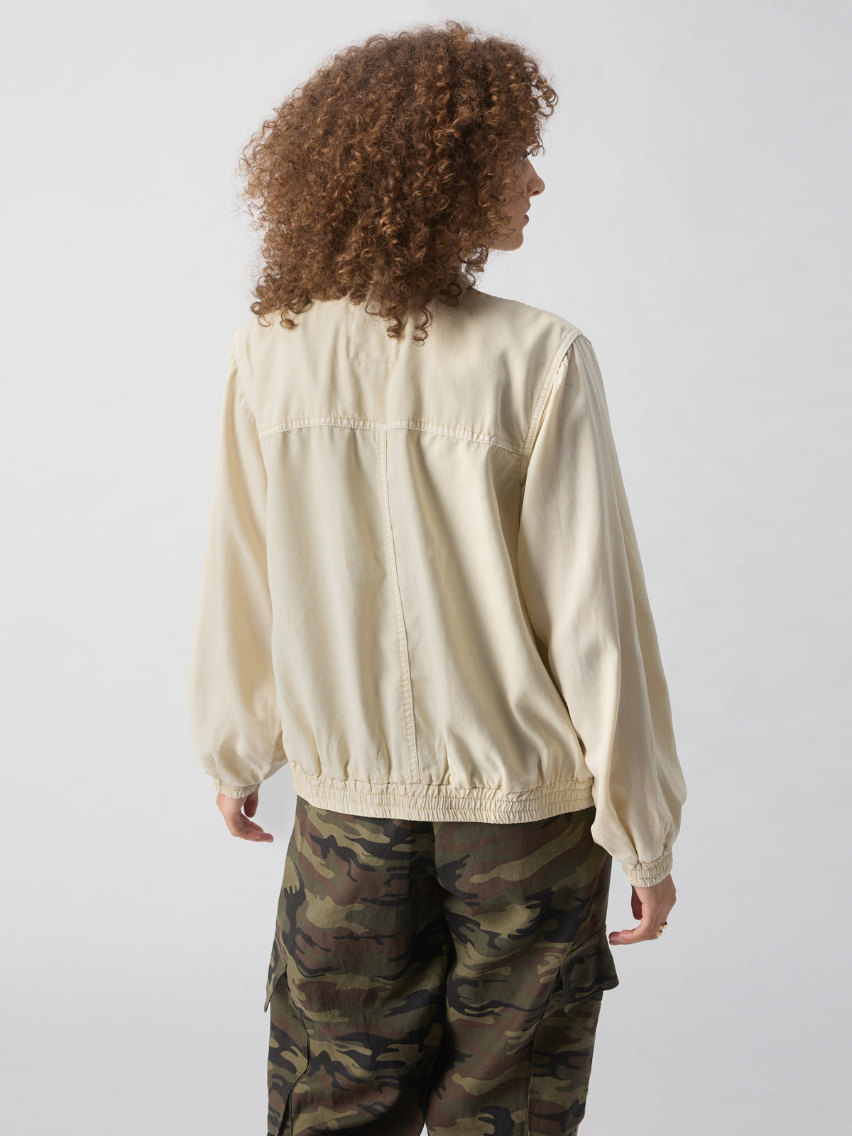 A person with curly hair, wearing the Field Utility Jacket Birch from Sanctuary Clothing and green camouflage pants, stands facing away from the camera against a plain background.