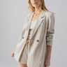 A person with long, blonde hair is wearing the Sanctuary Clothing Refresh Blazer Vineyard Stripe over a white top, paired with matching beige shorts. They are looking into the camera with a neutral expression, hands relaxed by their sides, standing against a plain, light grey background.