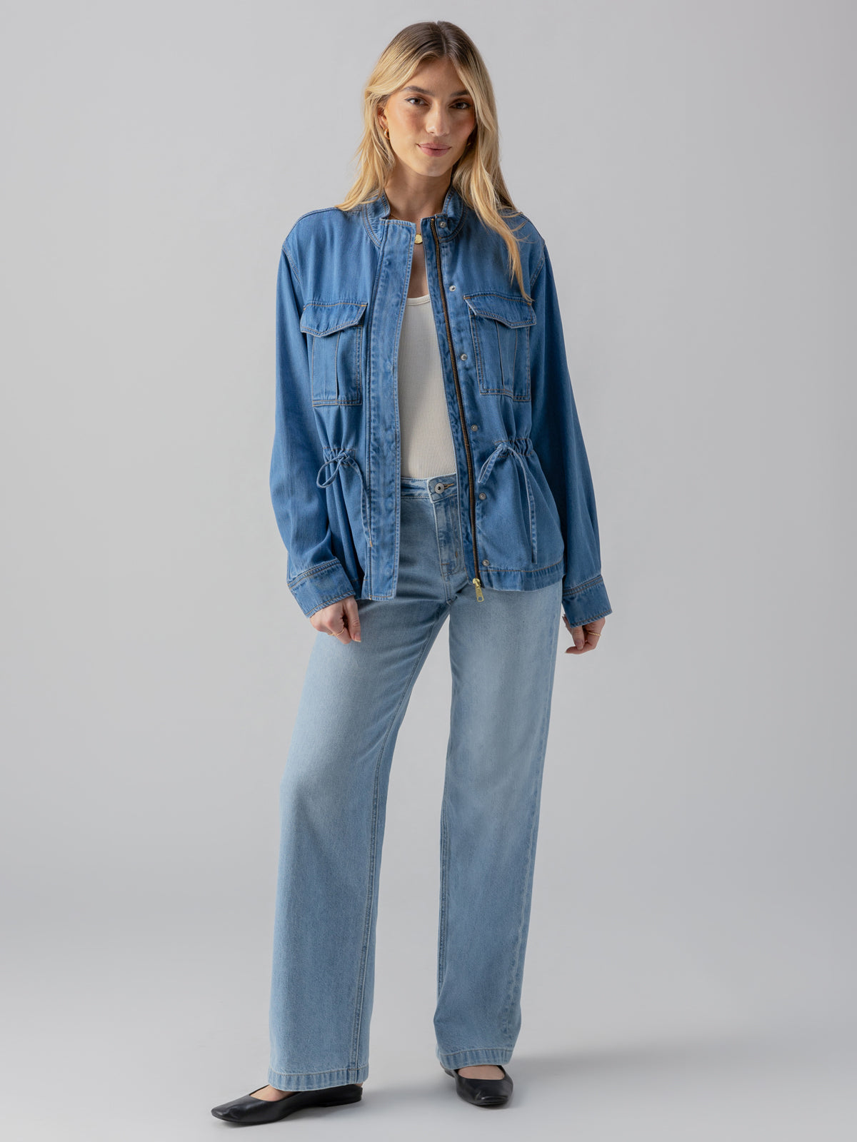 A woman with long, blonde hair is standing against a plain background. She is wearing the Cinched Surplus Jacket Sun Drenched by Sanctuary Clothing over a white top, accompanied by light blue high-waisted jeans and black shoes. She has a relaxed and casual posture.