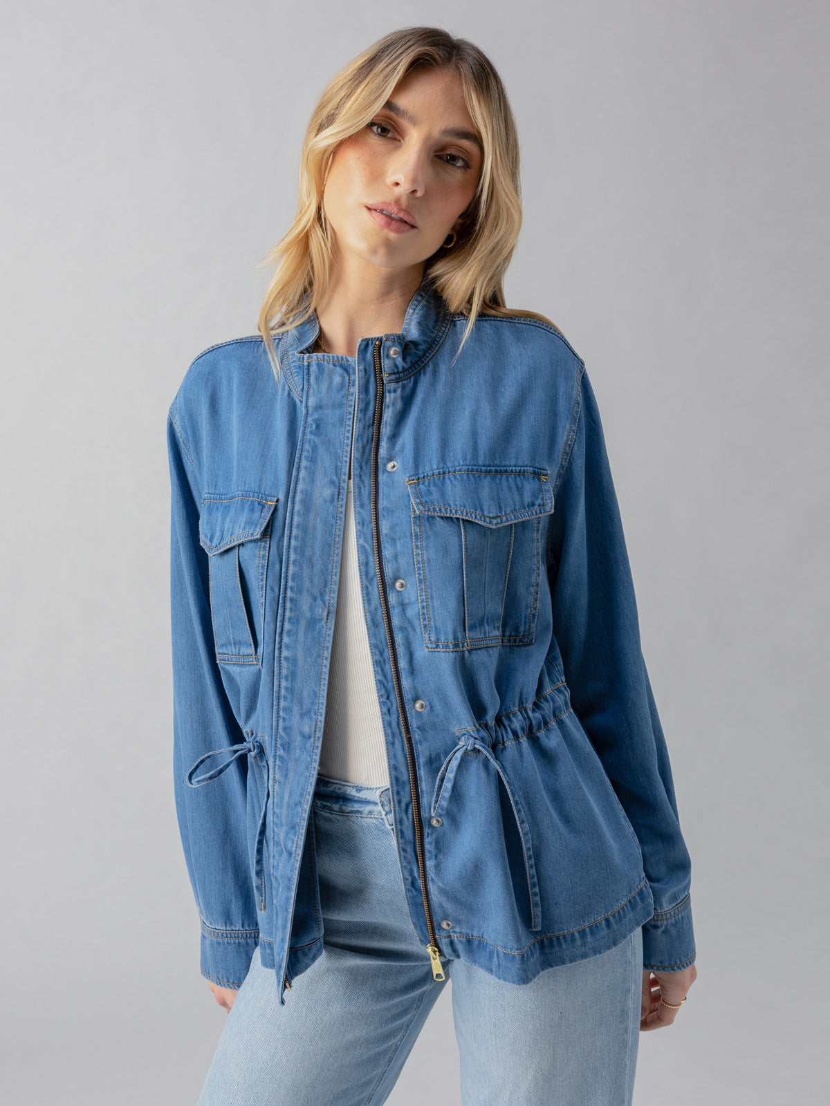 A person with light hair poses against a neutral background, donning the Sanctuary Clothing Cinched Surplus Jacket in Sun Drenched. The blue denim jacket features a front zipper, multiple pockets, and a drawstring waist, worn over a white top. They are looking slightly to the right with a relaxed expression.