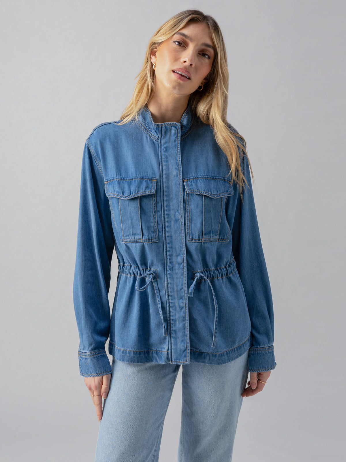 A woman with long blonde hair is wearing the Cinched Surplus Jacket Sun Drenched by Sanctuary Clothing. The blue denim jacket features front pockets and drawstring detailing at the waist. She is also wearing light blue jeans. She stands against a plain gray background, facing the camera, with a neutral facial expression.