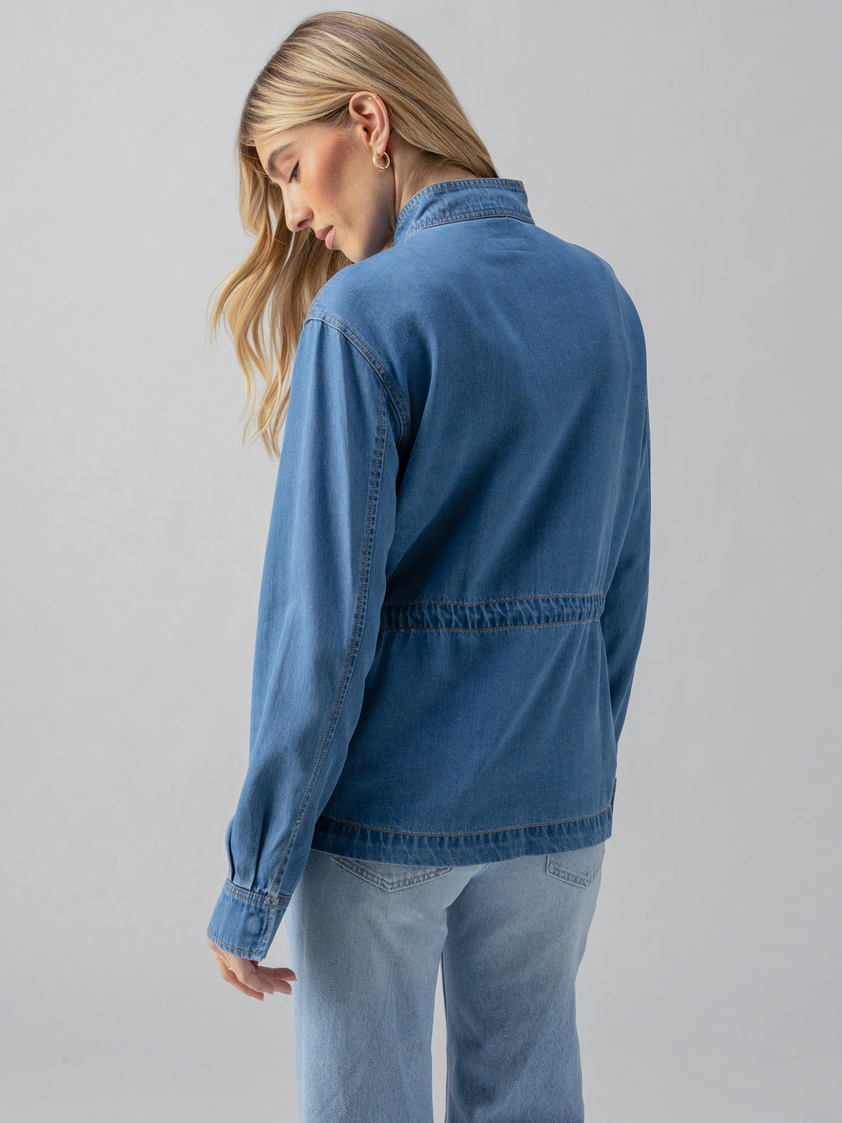 A person with long, blonde hair is shown from the back, wearing a Sun Drenched Cinched Surplus Jacket from Sanctuary Clothing and light blue jeans. They are standing against a plain grey background.