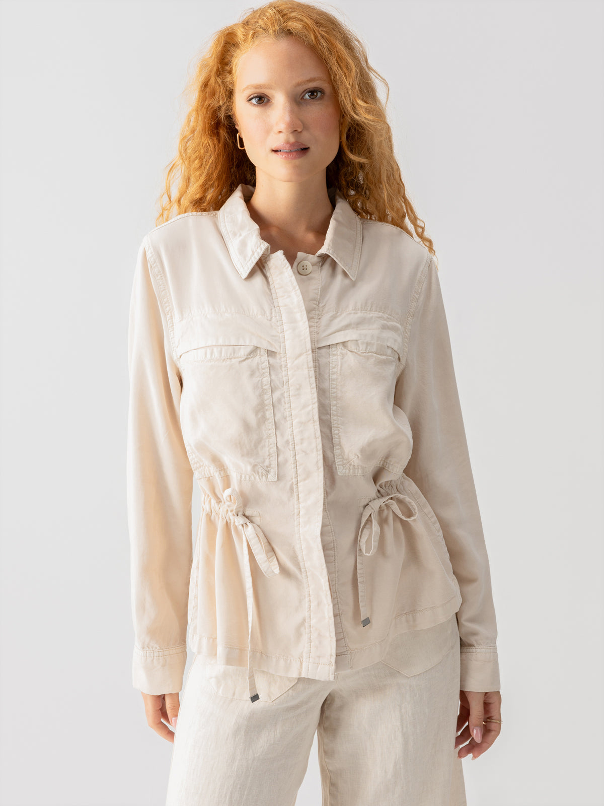 A woman with wavy, red hair is wearing a Lora Surplus Jacket Crystal Stone by Sanctuary Clothing, featuring button-up details and a drawstring waist. She pairs it with matching light beige pants, standing against a plain white background while looking directly at the camera.