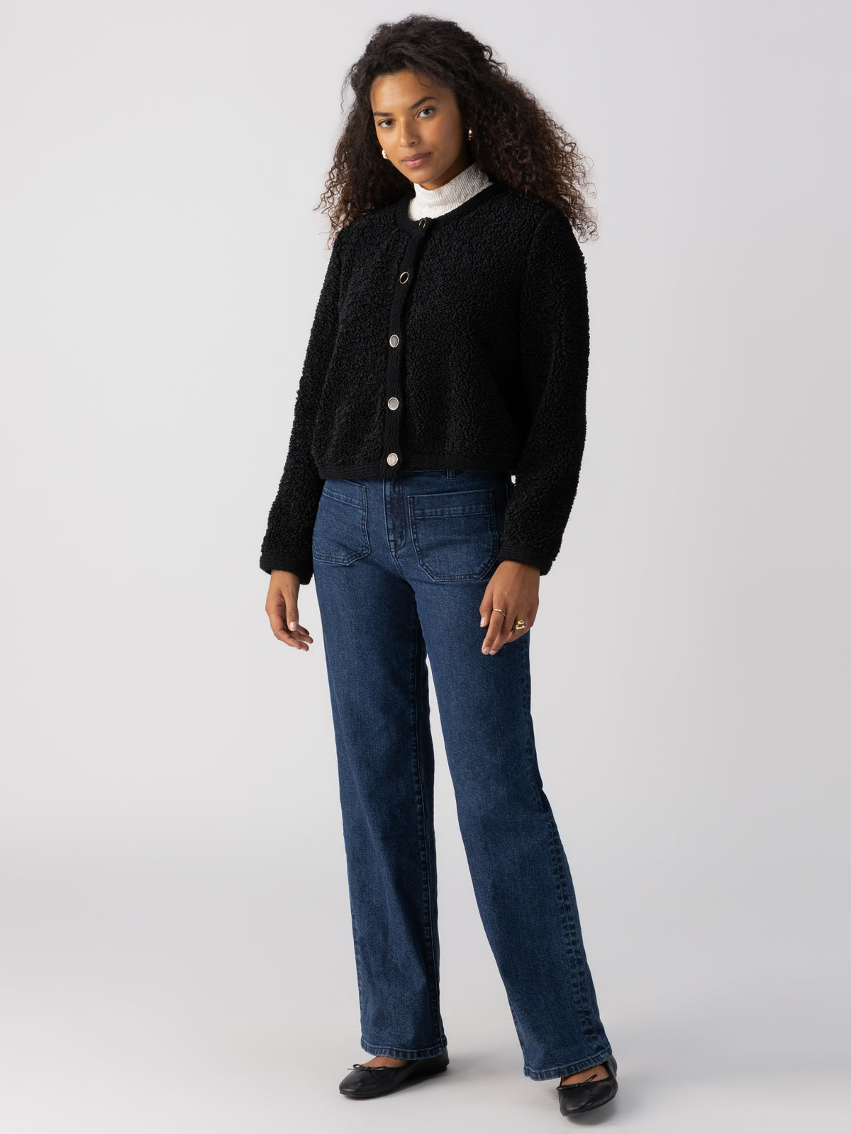 A person with curly hair stands against a plain background, wearing the Cozy Cardigan Black by Sanctuary Clothing over a white turtleneck. They also have on blue jeans and black shoes. Their hands are relaxed by their sides.