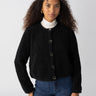 A person with long, curly hair is wearing the Cozy Cardigan Black by Sanctuary Clothing over a white turtleneck and blue jeans. They are standing in front of a plain, light-colored background, looking directly at the camera with a slight smile.