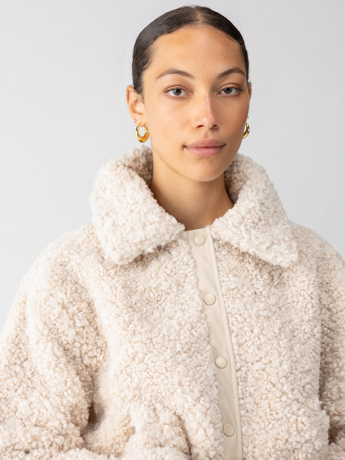 A person with sleek, tied-back hair is wearing the Libby Sherpa Jacket in Toasted Almond by Sanctuary Clothing; the plush, cream-colored textured coat features a large collar and button-up front. They accessorize with gold hoop earrings and stand against a plain, light grey background.